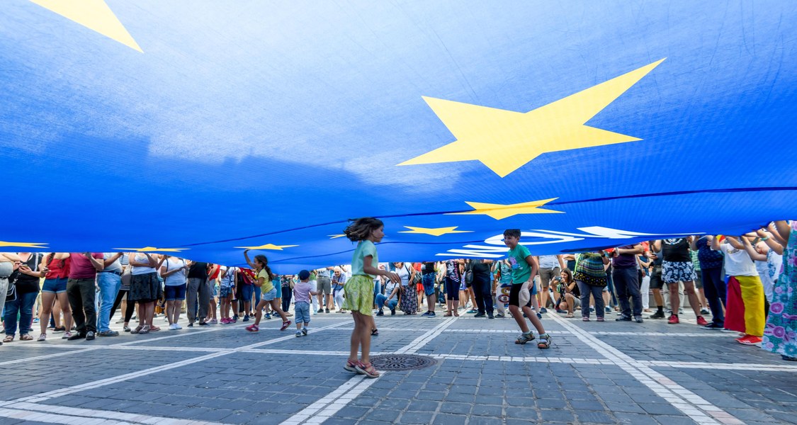 Kids are playing under a huge flag of the EU. The Flag is held up on all sides by a lot of grown-ups.