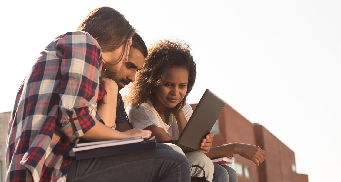 Three young adults sitting next to each other outside, reading something from one laptop.