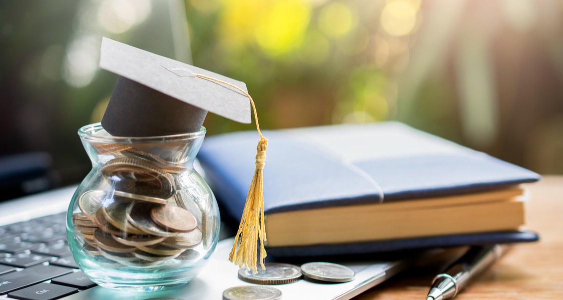 A jar full of coins on a table with a book and a laptop.