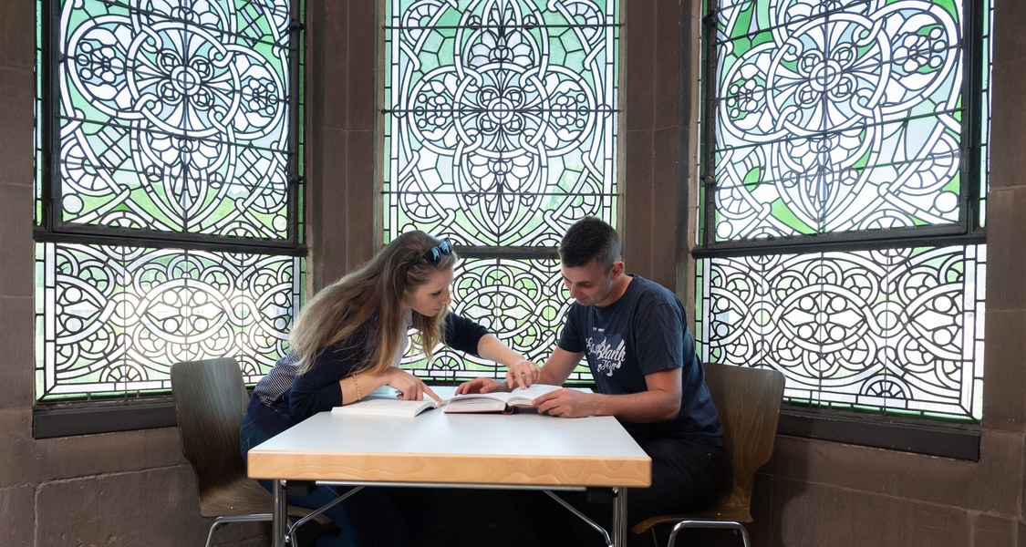 Two students sit in front of an ornate window and study together.