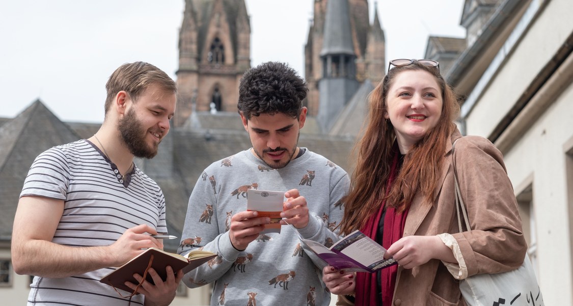 Three students are in conversation in the foreground as they look at a brochure. The background shows the peaks of a church.